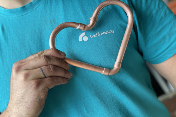 Ein Mitarbeiter der Firma hält ein frisch aus Kupferrohr gelötetes Herz vor das bad-und-heizung-Logo