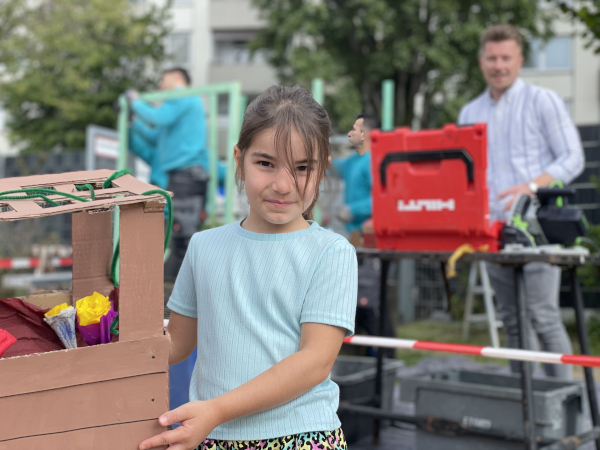 Kindergartenkind hält im Vordergrund ein Modell der Rankhilfe. Im Hintergrund steht Sebastian Söll von Sanitherm neben einem Hilti-Koffer, während seine Mitarbeiter die Rankhilfe für die Kindertagesstätte bauen.